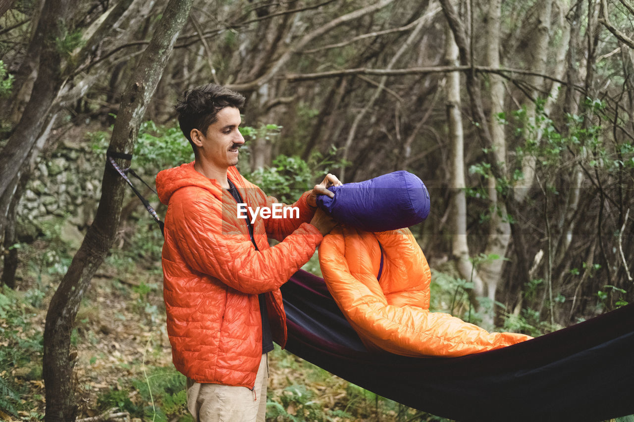 A man packs a sleeping bag and hammock at campsite in the forest