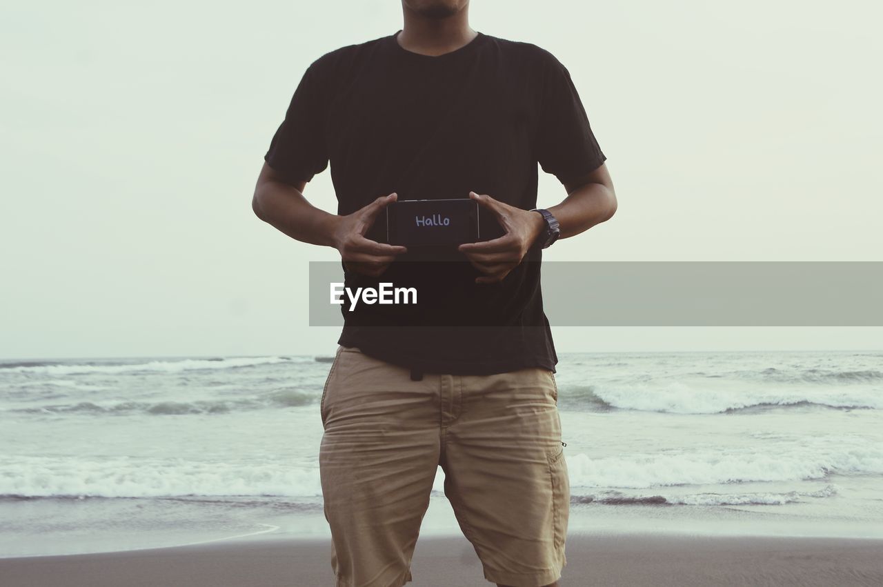 Midsection of man showing message on smart phone standing at beach against sky