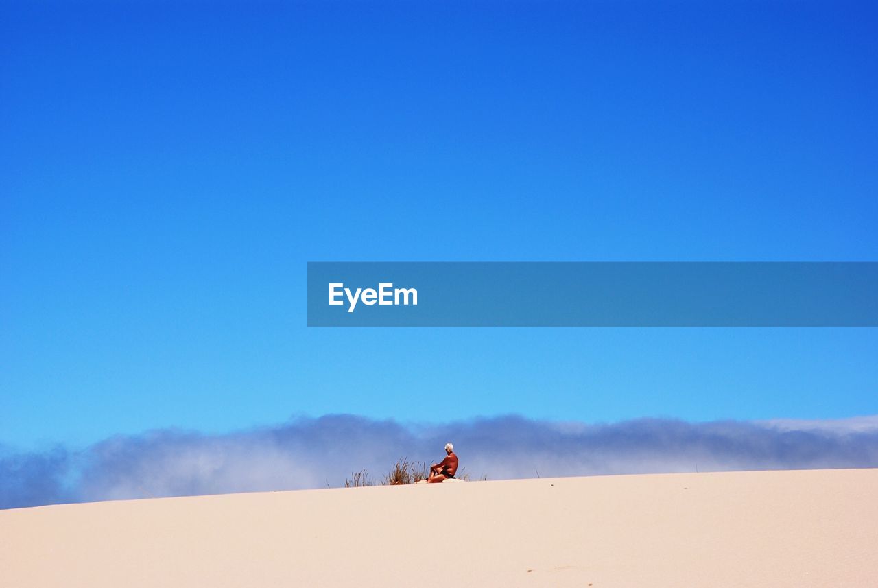 Shirtless man sitting on sand dune at desert against blue sky