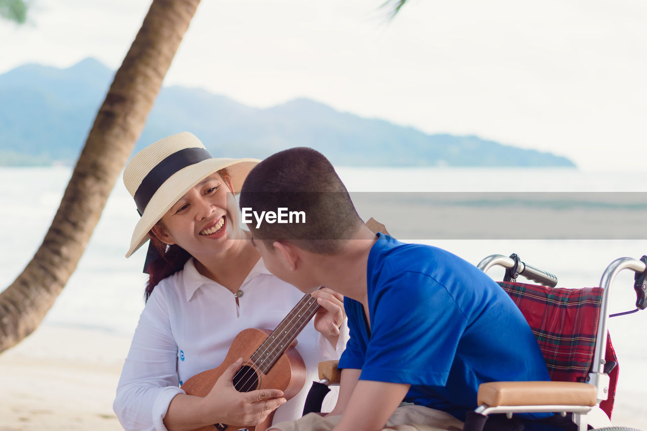 Smiling mother playing ukulele while sitting with son at beach