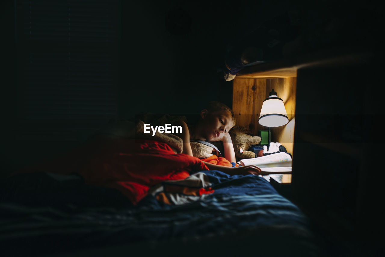 Boy reading book in darkroom under electric lamp at home