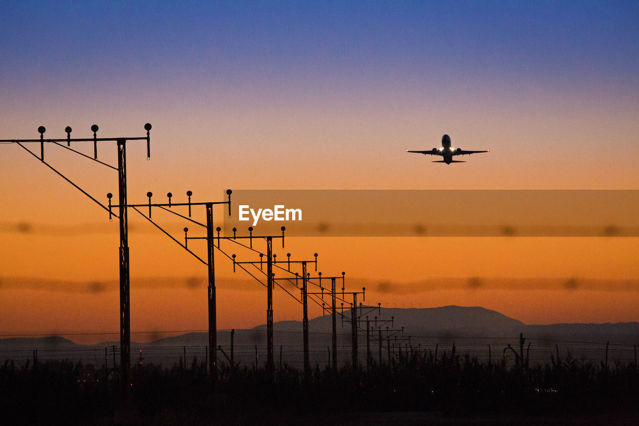 Poles on field against silhouette airplane flying in sky during sunset