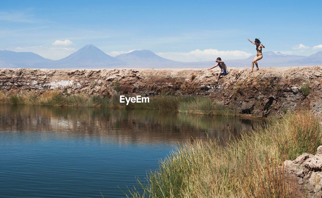 Scenic view of mountains against sky and a young man and woman jumping to a lake 