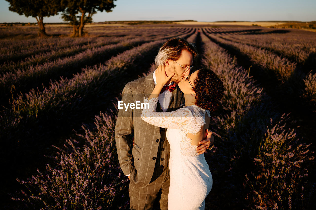 High angle side view of romantic newlywed couple standing face to face kissing on spacious field against purple sunset sky