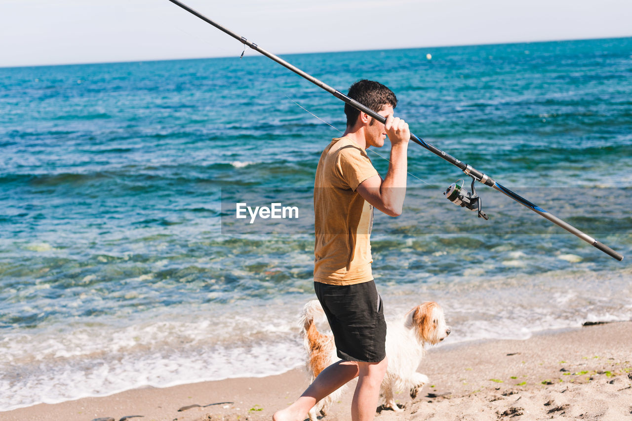 Man holding fishing rod while walking with dog at beach