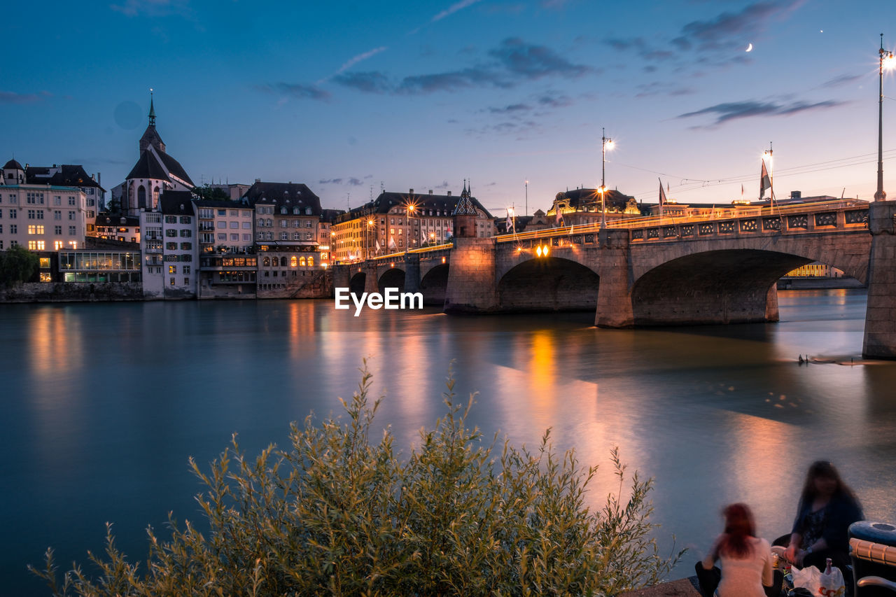 Friends sitting near illuminated bridge over river in city at dusk