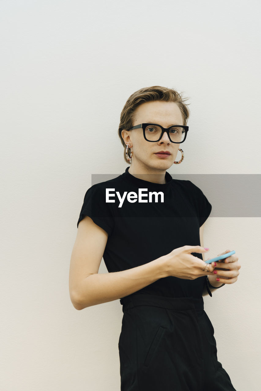 Confident androgynous entrepreneur standing with smart phone against while wall in board room