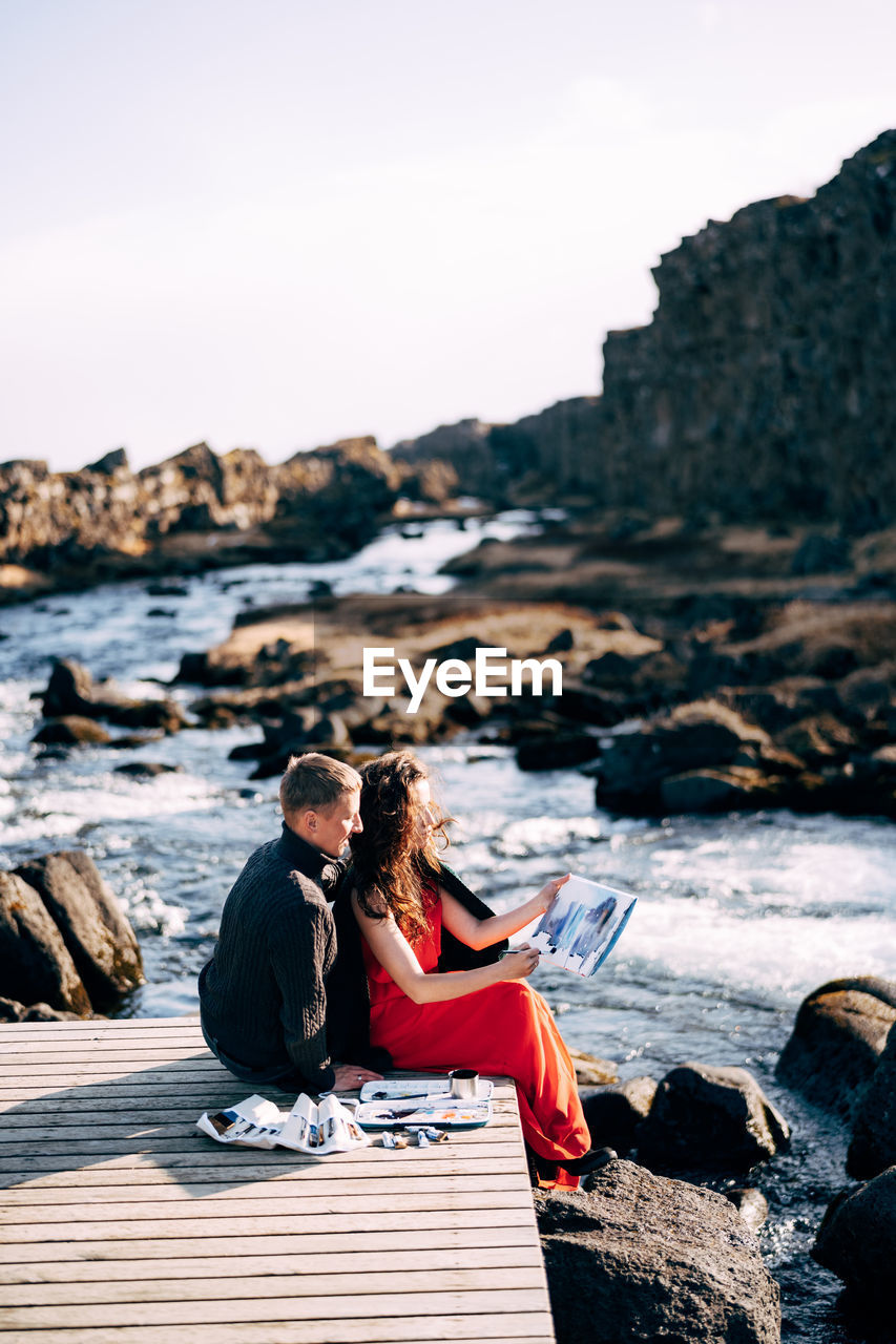 People sitting on rock by sea against sky