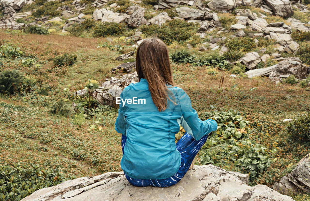 Rear view of meditating young woman in blue sitting on stone in green mountain valley,  mindfulness