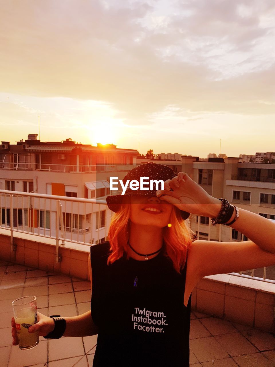 Woman holding drink while standing on terrace in city against sky