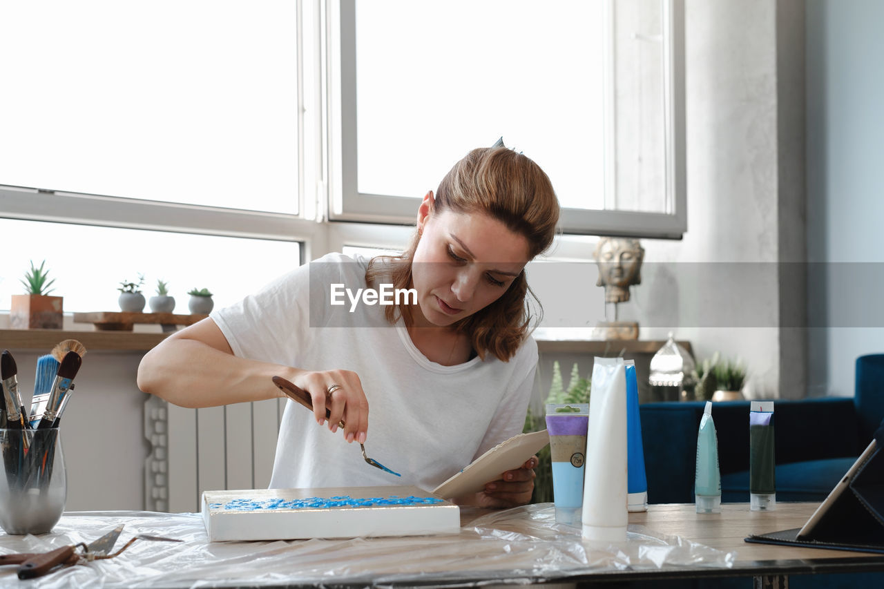 Woman artist painting picture at home with acrylic paints and spatula over the window. adult hobby