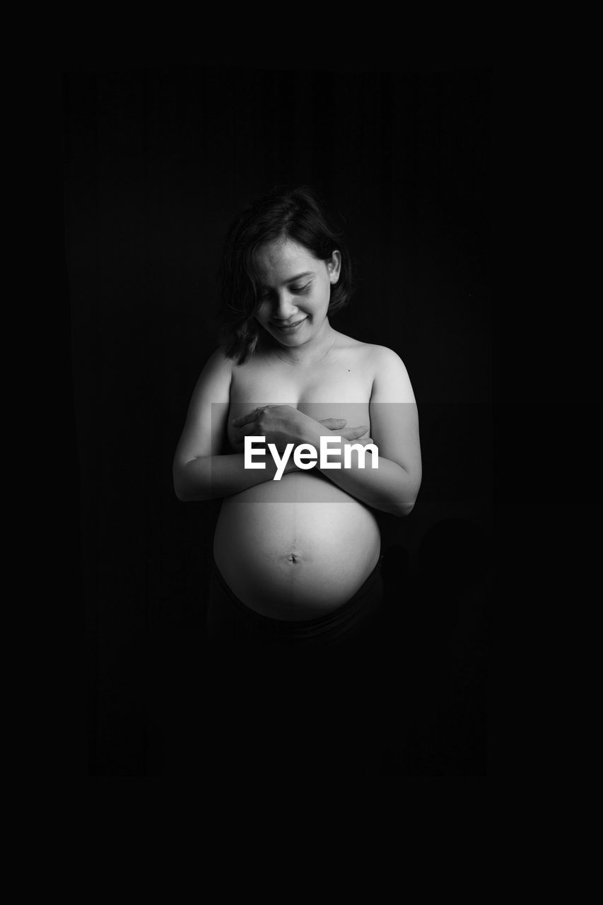 Topless pregnant young woman standing against black background