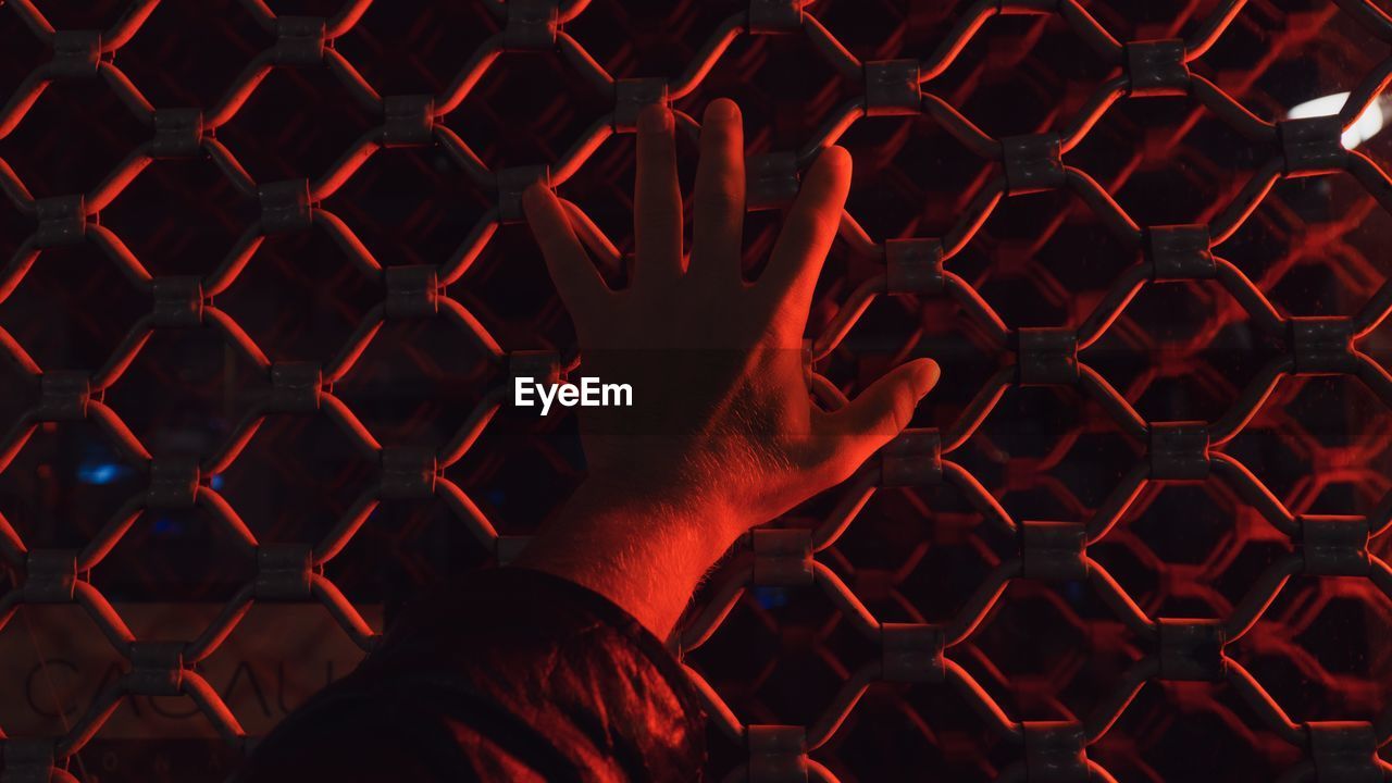 Cropped hand of man touching fence with illuminated red light at night