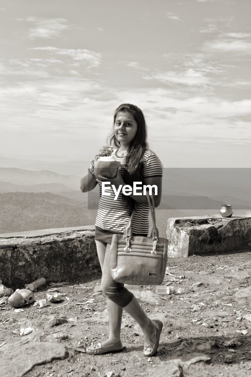 Portrait of smiling young woman drinking coconut water while standing against sky