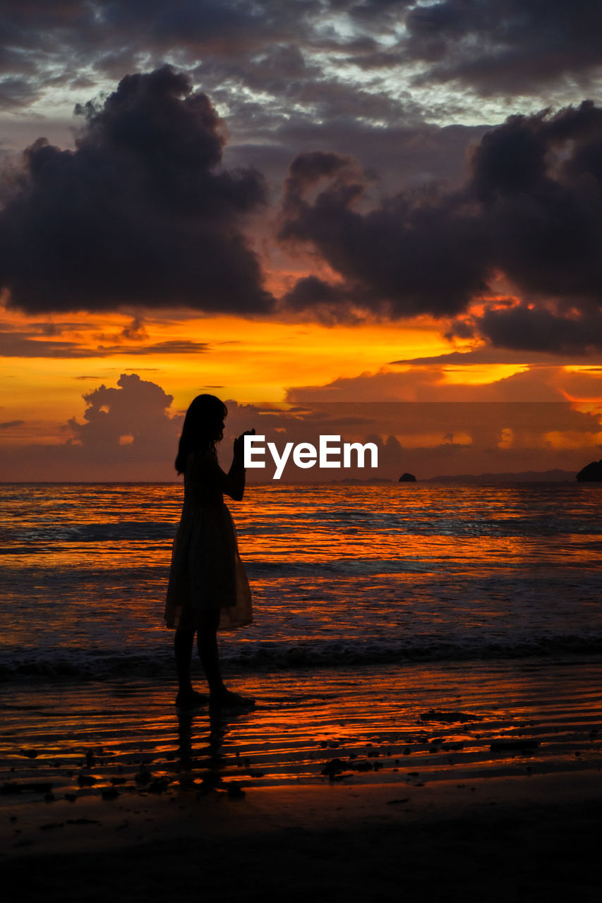 Silhouette woman praying while standing on beach at dusk