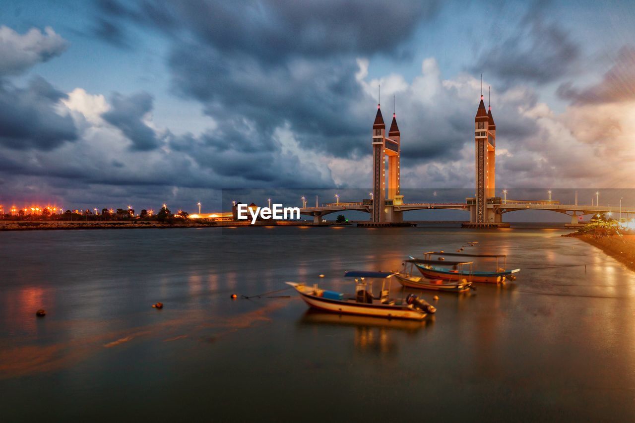 Boats moored in river against dramatic sky and bridge