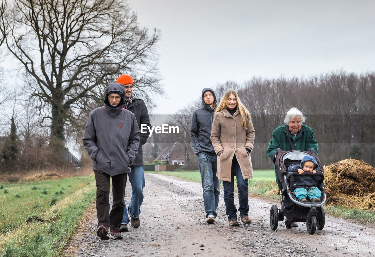 Family walking on dirt road by field against sky