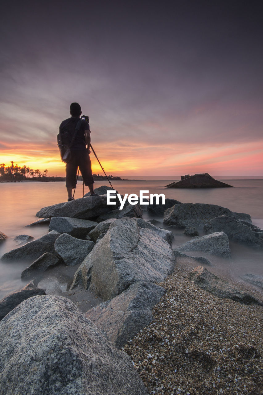 Man with tripod standing on rock at beach against sky during sunset