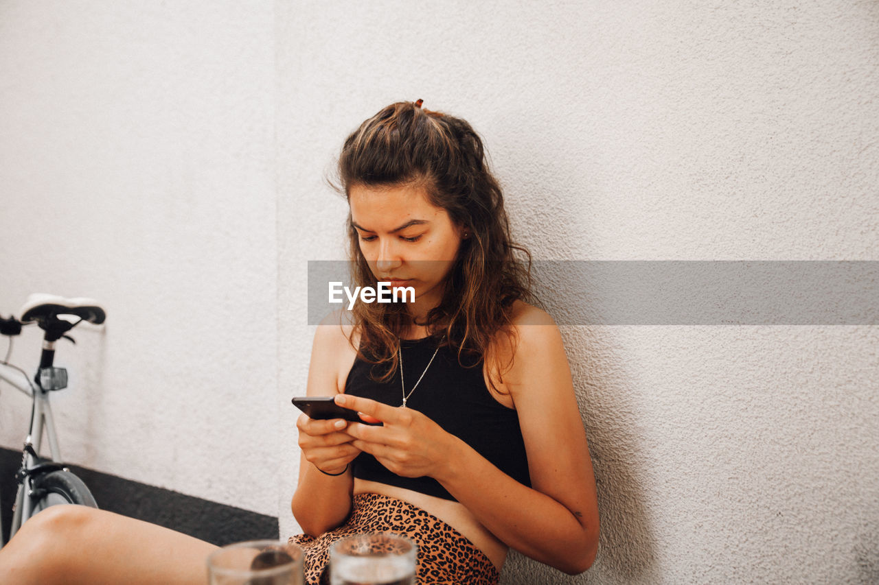 Young woman using mobile phone while sitting on wall