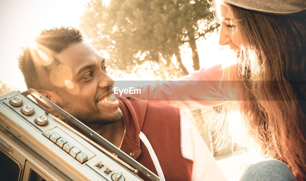 Close-up of cheerful young couple with radio against trees during sunset