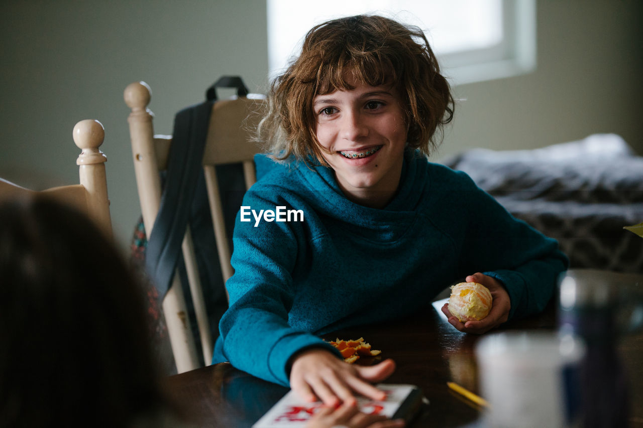 Girl with braces smiles at her sibling while holding an orange