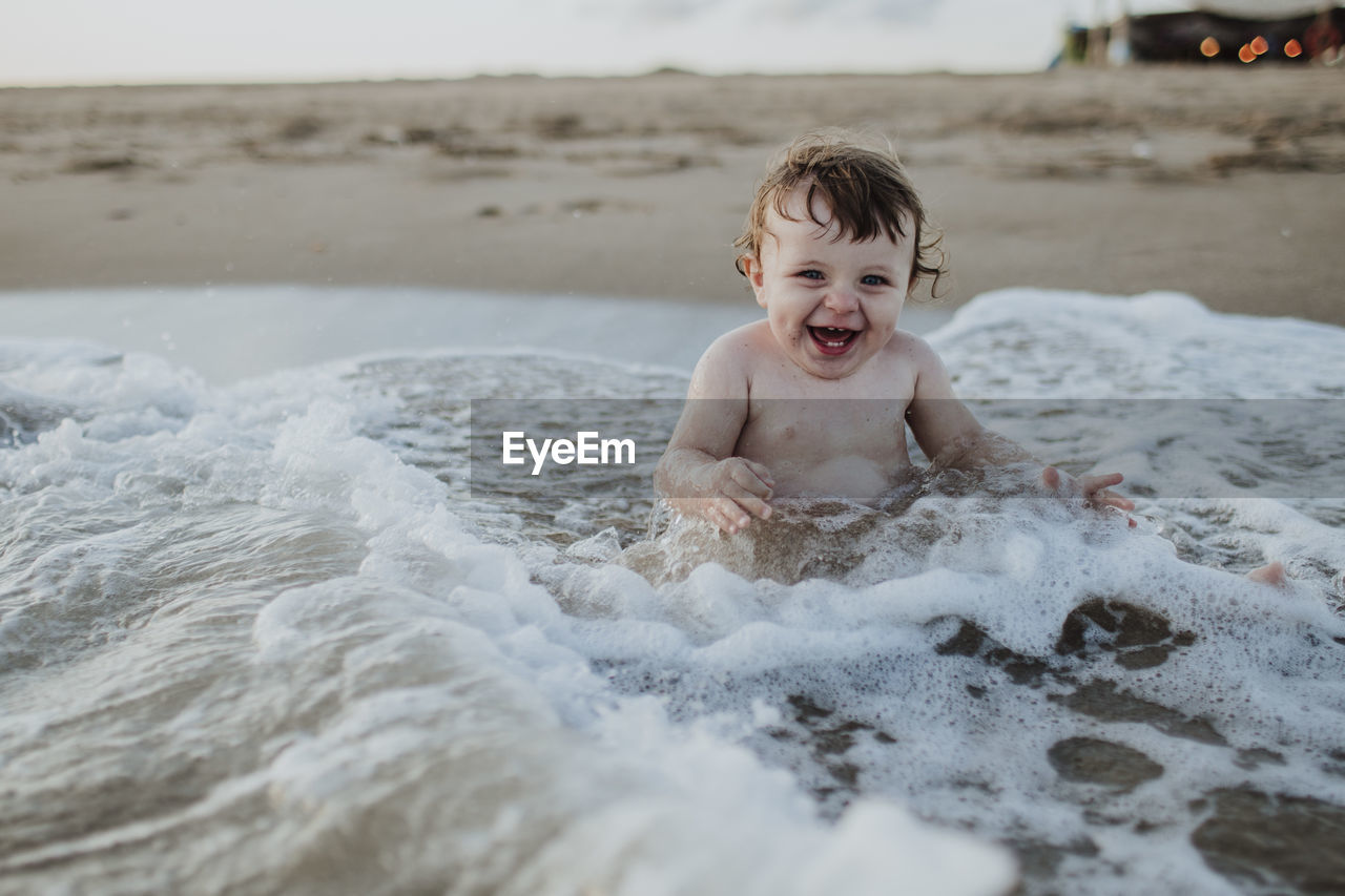 Male toddler laughing while sitting in water at beach during sunset