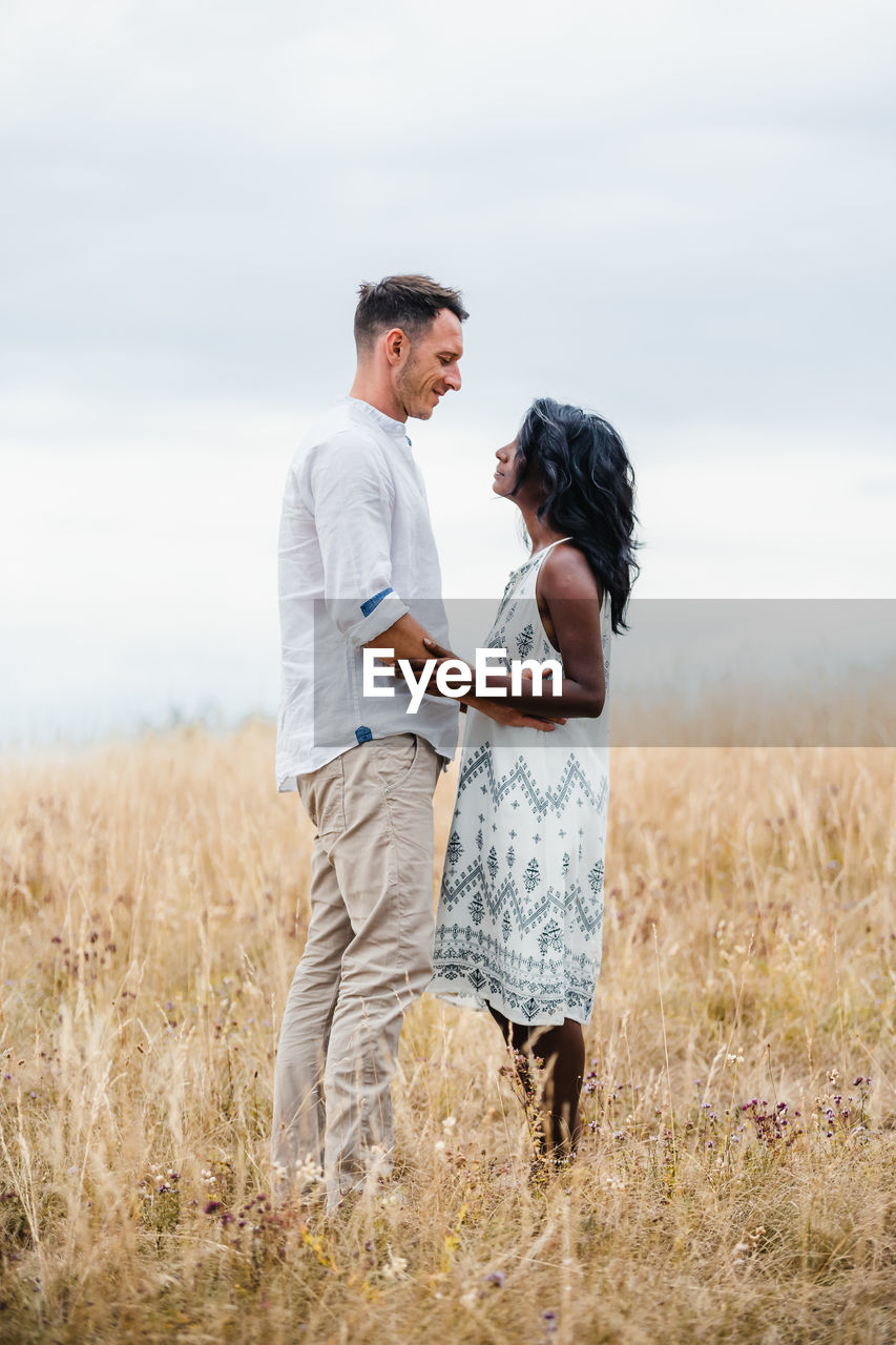 Side view of smiling man embracing indian girlfriend while looking at each other in field under cloudy sky