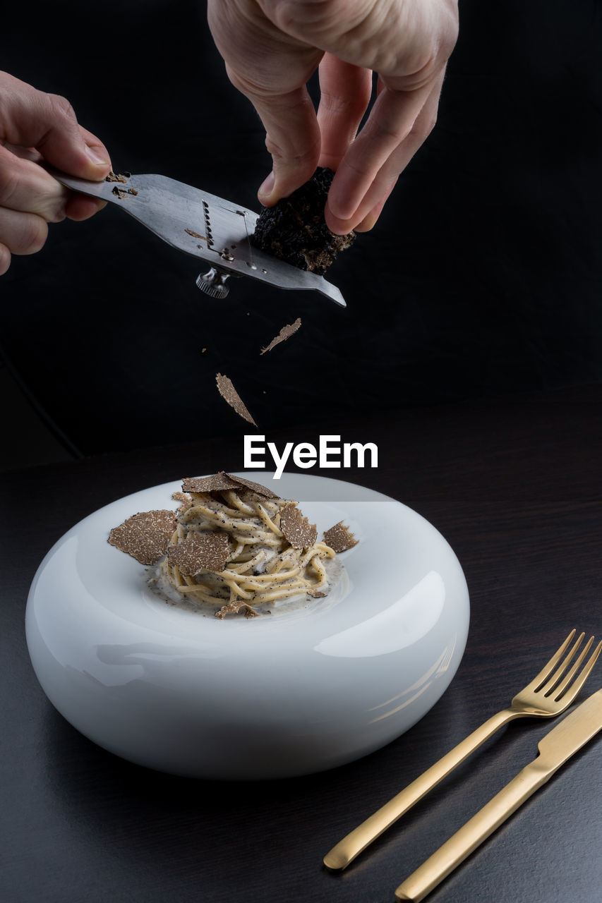 Hands of crop anonymous chef grating delicious black truffles on pasta served in white plate near cutlery in modern restaurant