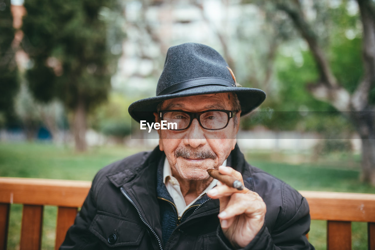 Happy senior male with mustache wearing hat smoking cigar and looking at camera while sitting on bench in park with green trees on blurred background