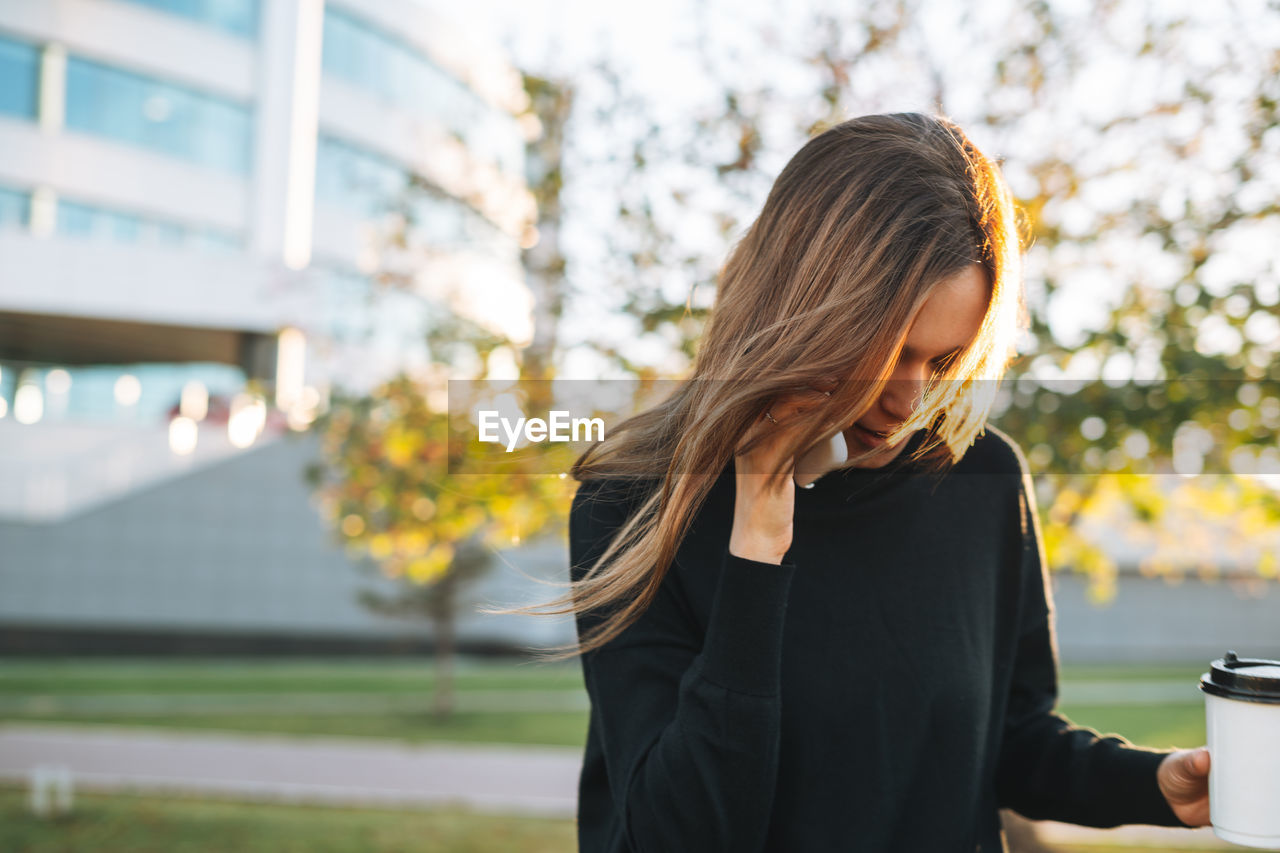 Portrait of young woman student with long hair using mobile phone in city park in golden hour