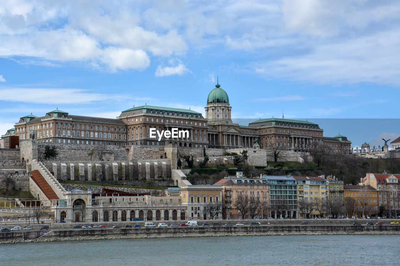 Danube river by buildings and buda castle against cloudy sky