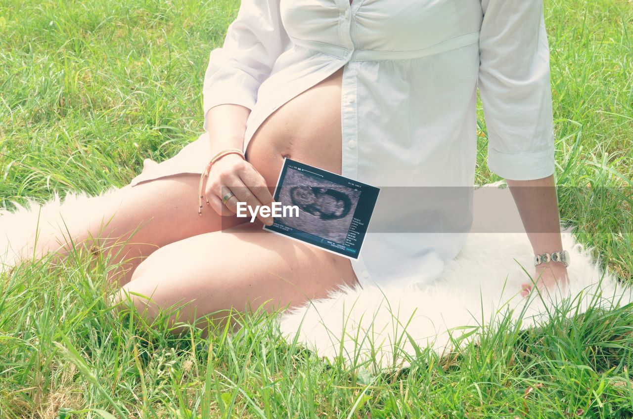 Midsection of pregnant woman holding ultrasound photograph while sitting on grassy field