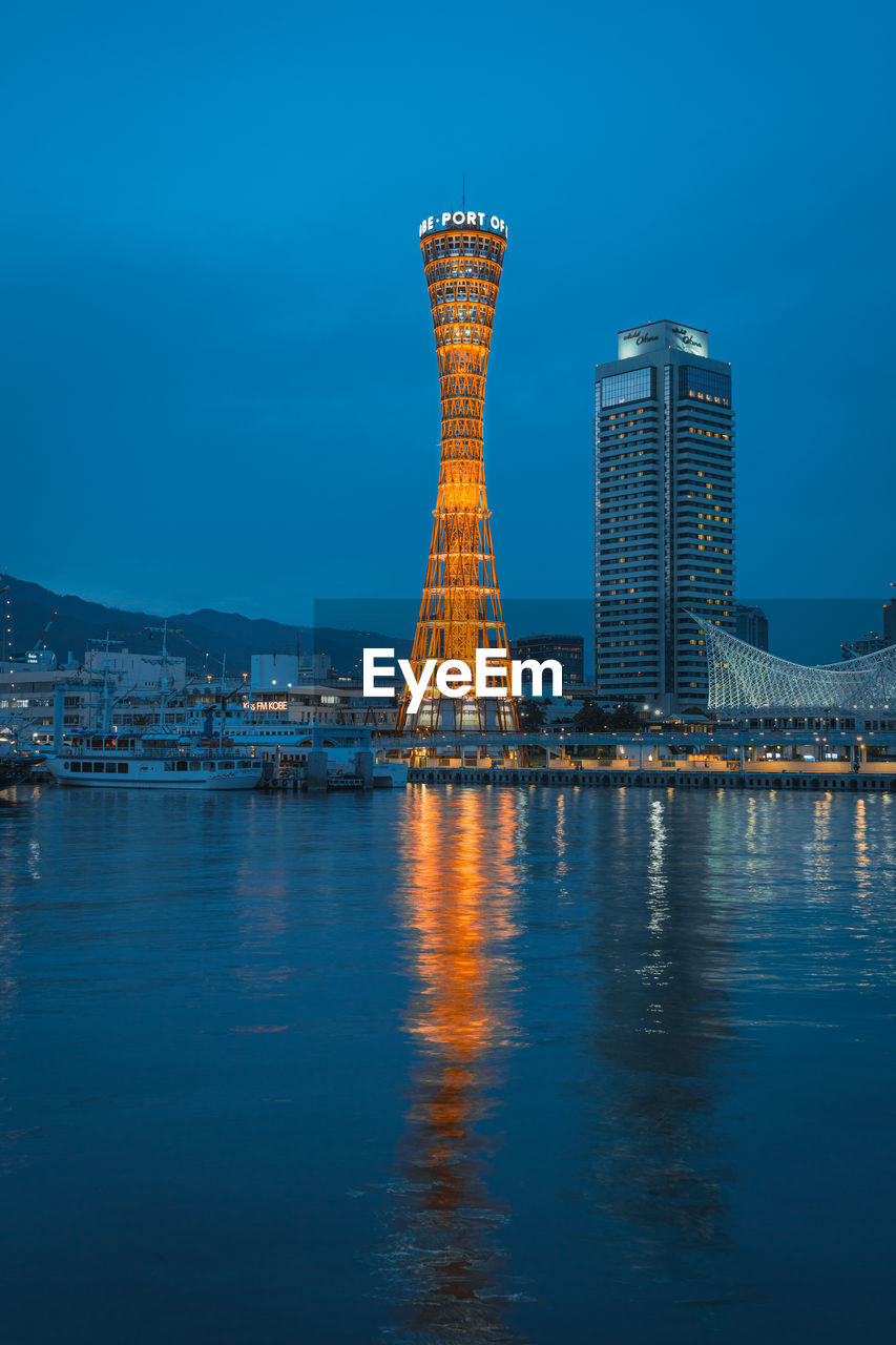 ILLUMINATED BUILDINGS BY SEA AGAINST BLUE SKY AT DUSK