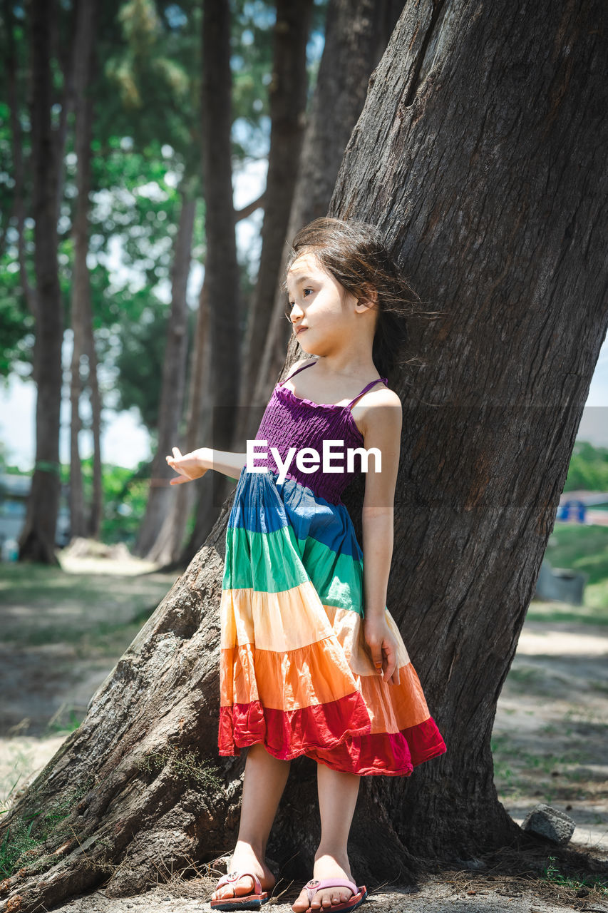 Pretty little girl wearing colourful dress and standing near the beach trees.