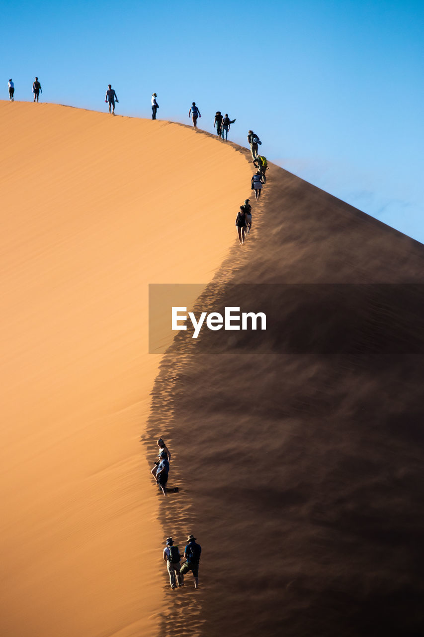 People walking on sand dune against clear blue sky