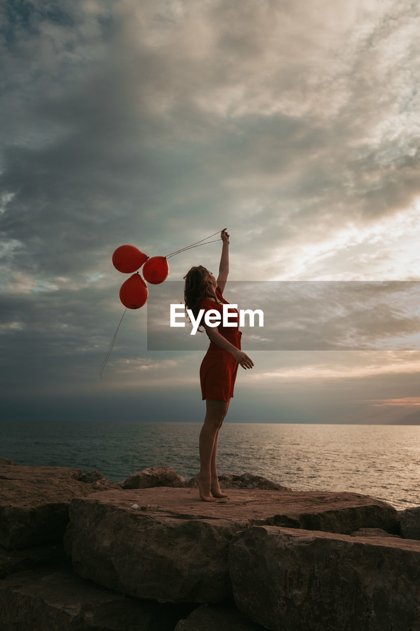 Woman holding balloons while standing on rock by sea against sky during sunset