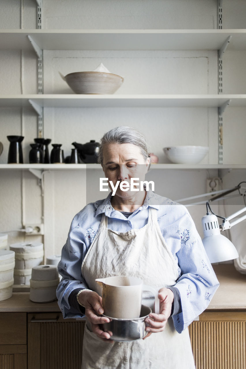 Senior female potter standing with pitcher against shelves at workshop