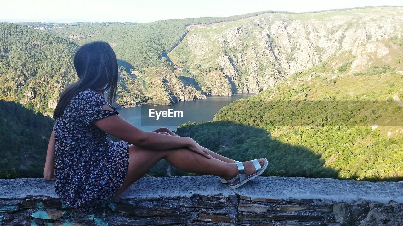 Full length of young woman sitting on retaining wall by lake and mountains