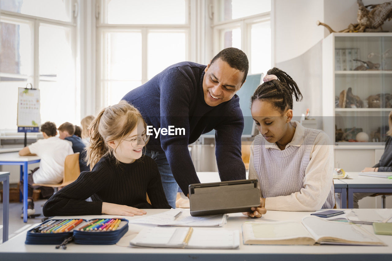 Smiling male teacher doing e-learning with female students in classroom