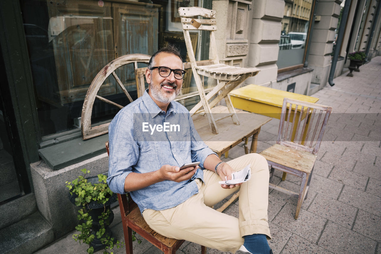 Portrait of owner holding mobile phone while sitting outside antique shop