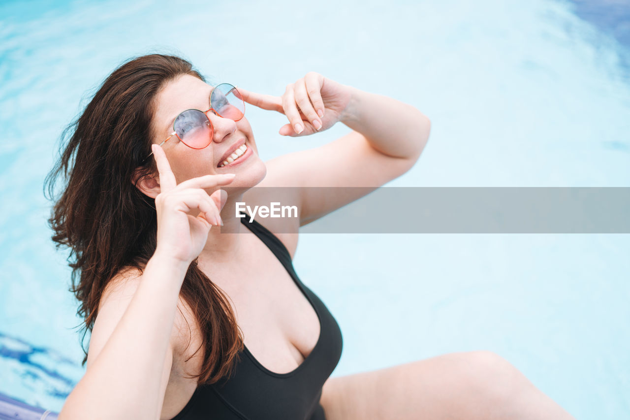 Stylish happy young woman plus size body positive in black swimsuit and sunglasses near hotel pool