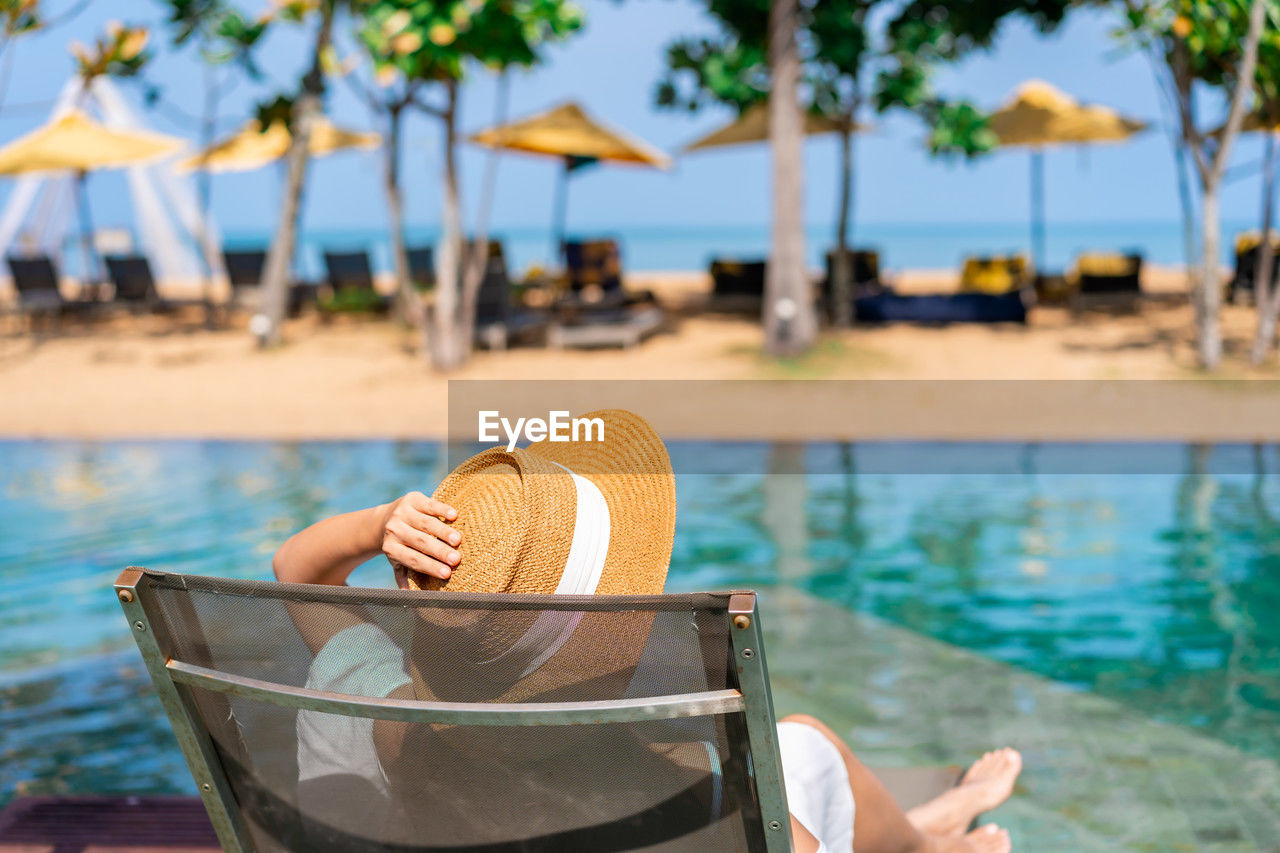 water, relaxation, vacation, trip, holiday, nature, summer, one person, leisure activity, tropical climate, swimming pool, chair, sea, adult, travel destinations, tourist resort, tranquility, beach, lounge chair, travel, day, land, sunlight, focus on foreground, tranquil scene, tree, lifestyles, women, outdoors, palm tree, idyllic, sky, hat, sunbathing, food and drink, beauty in nature, resting, sitting, tourism, sun tanning, poolside, weekend activities, enjoyment, clothing, lying down, seat, plant, wealth, luxury, comfortable, sun, refreshment, tourist, young adult, deck chair, hotel, sunny