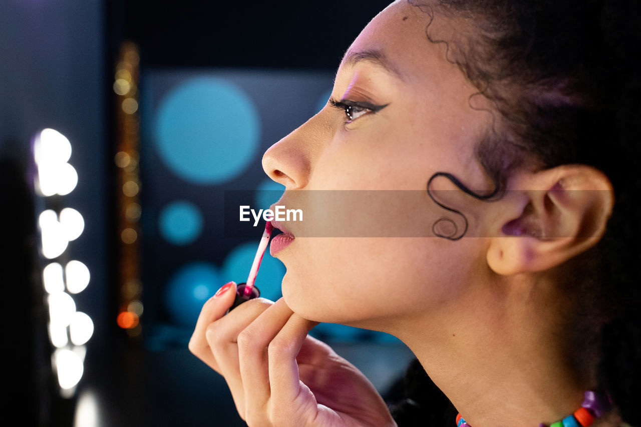 Side view of african american female applying lip gloss and doing makeup while looking in mirror with light bulbs in dark room