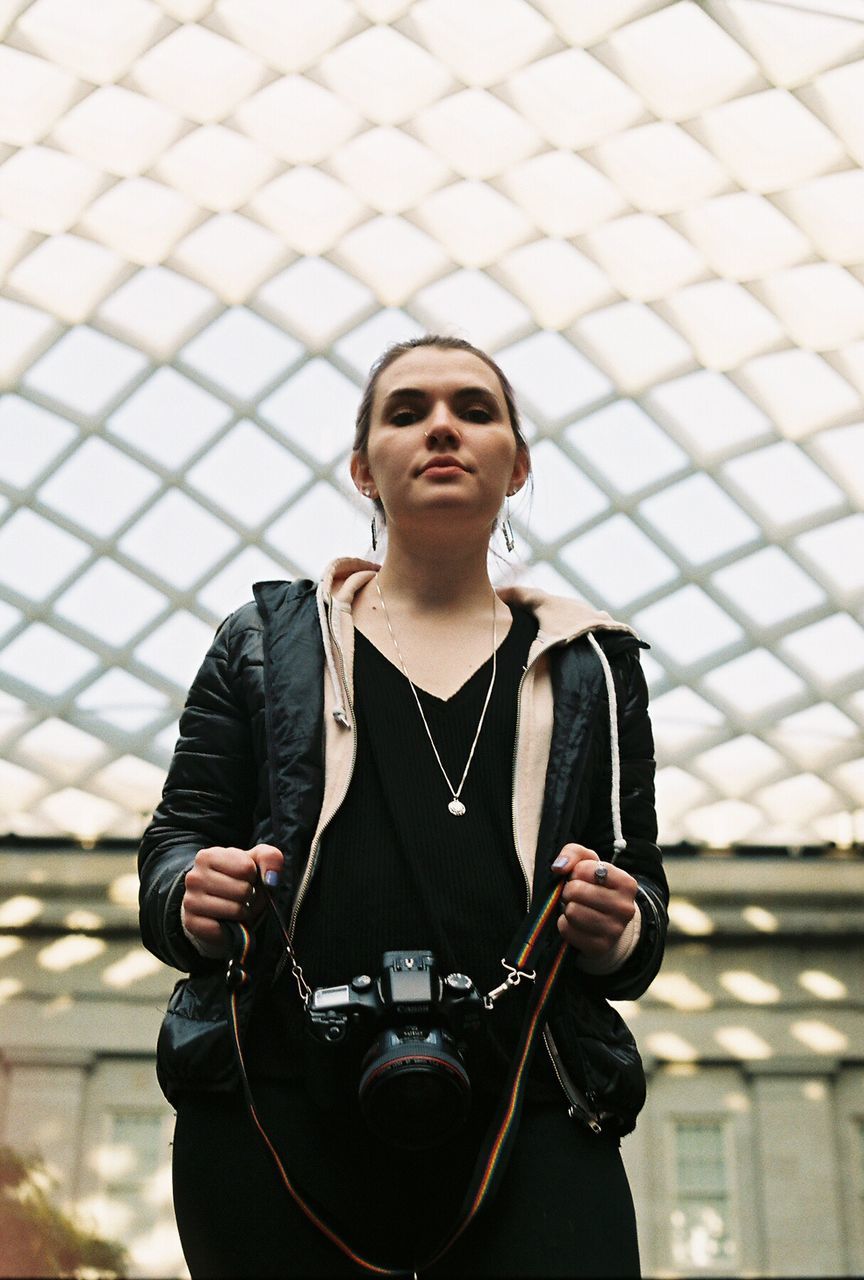 Low angle portrait of woman holding camera against skylight