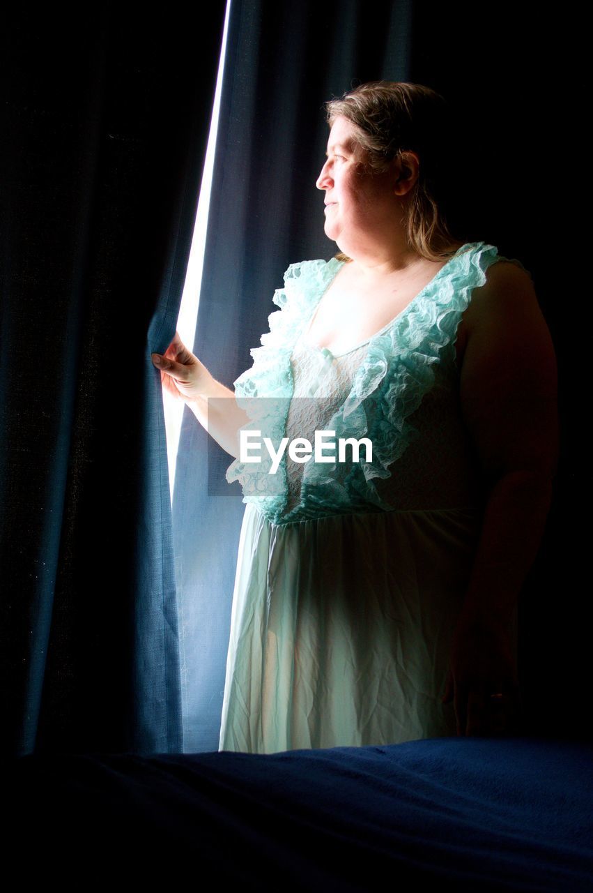 A beautiful middle-aged woman, in a lacy green nightgown, illuminated by window light.