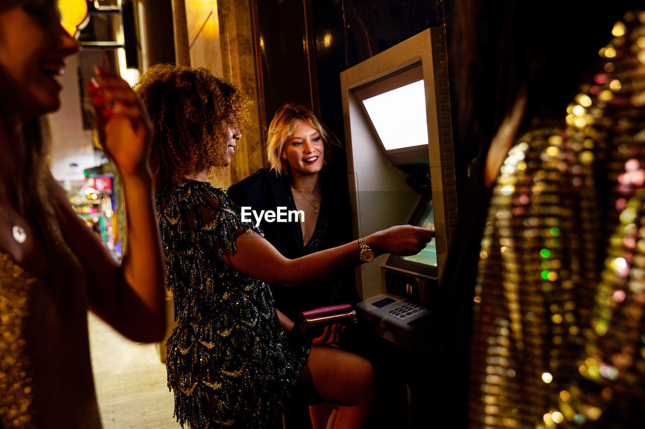 Smiling african american female inserting credit card and withdrawing cash from automated teller machine during party with friends