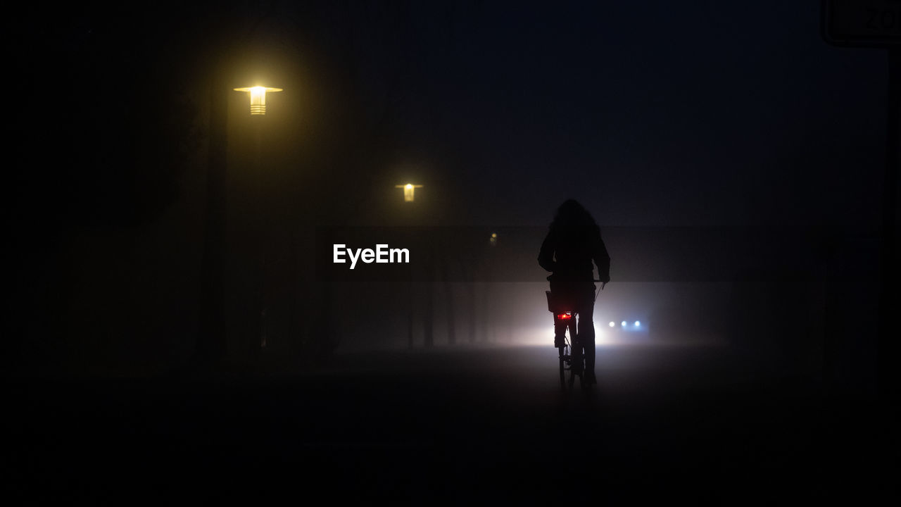 Rear view of dark silhouette person on bicycle on street at night