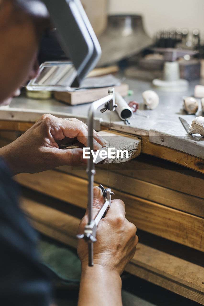 Cropped image of female artisan using handsaw while making jewelry on wooden table in workshop