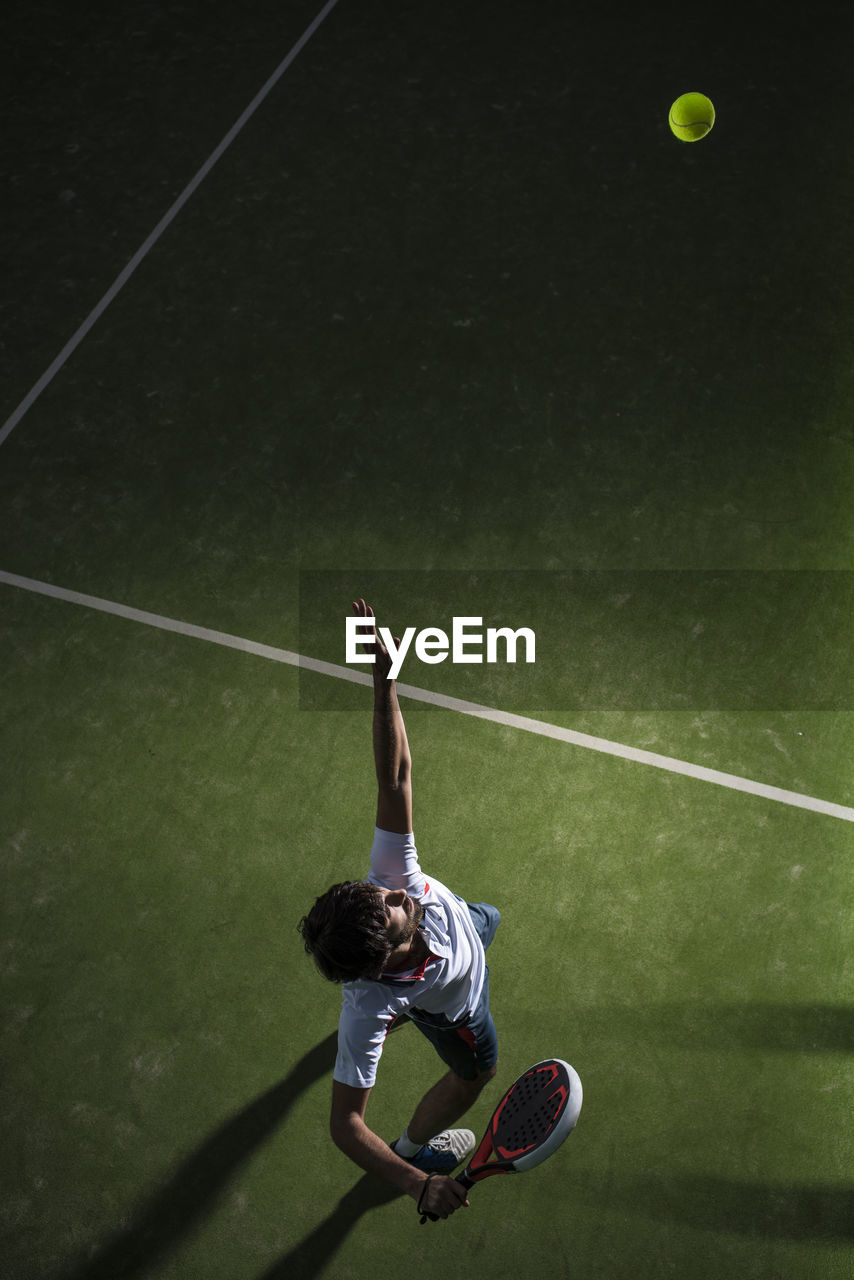 Male athlete servicing ball while playing padel tennis in sports court at night