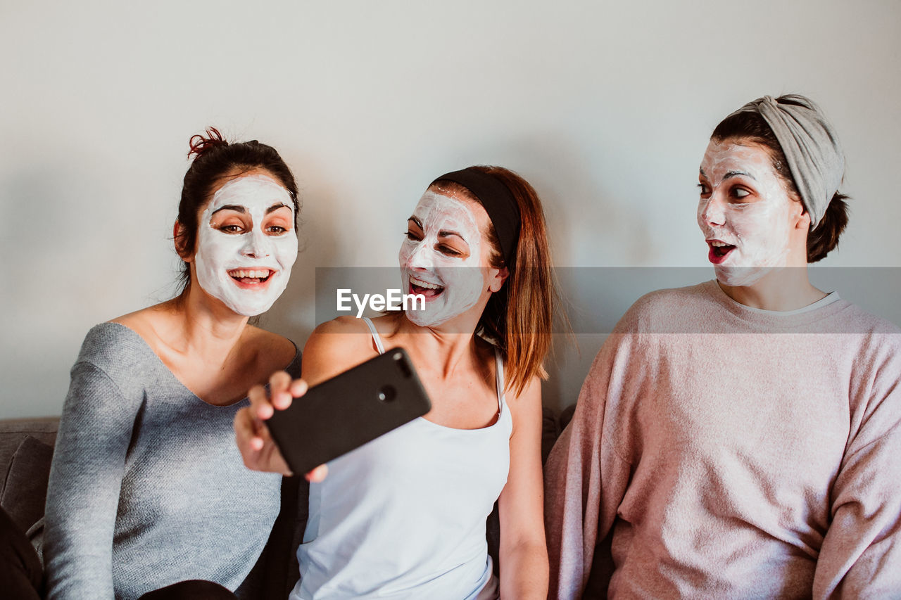 Female friends with facial masks taking selfie with against wall at home